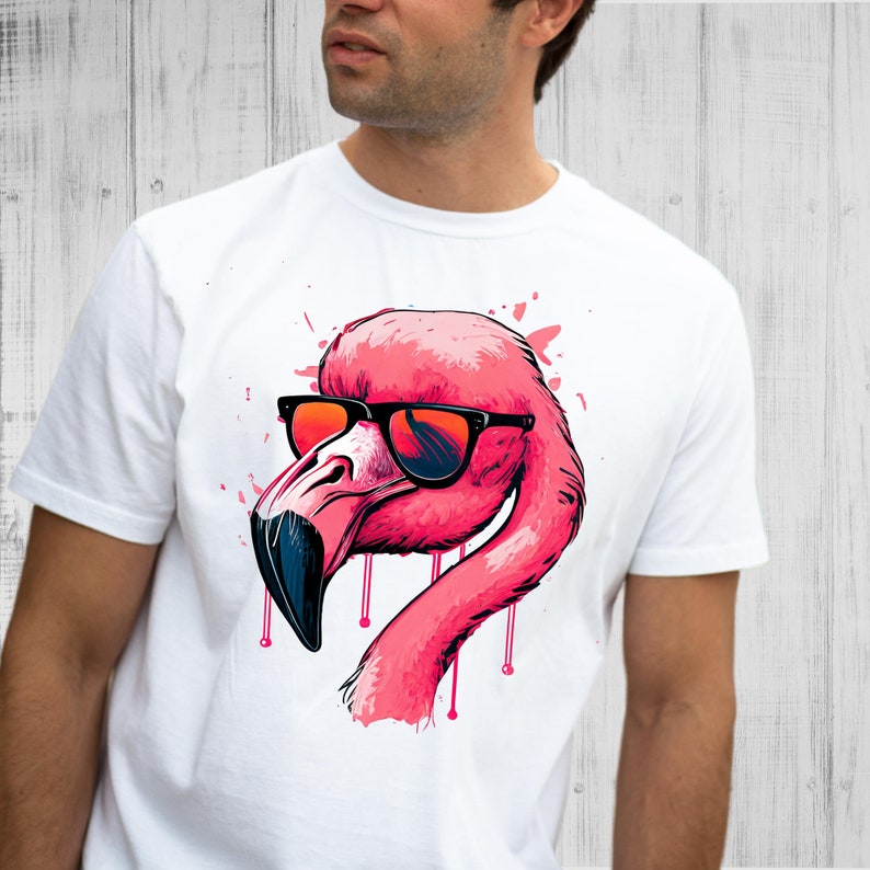 May include: White t-shirt featuring a pink flamingo wearing black sunglasses with orange lenses. The flamingo design has a stylized, cartoon-like appearance with pink paint splatter accents.