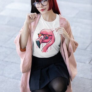 May include: White t-shirt featuring a pink flamingo wearing sunglasses. The flamingo design has a watercolor effect with red and pink accents. The shirt is worn by a person wearing glasses, a pink jacket, and a black skirt.