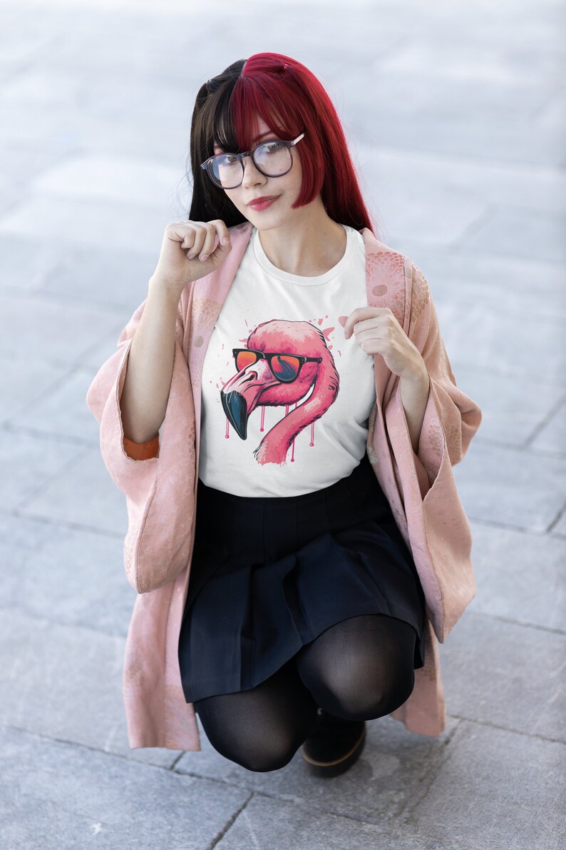 May include: White t-shirt featuring a pink flamingo wearing sunglasses. The flamingo design has a watercolor effect with red and pink accents. The shirt is worn by a person wearing glasses, a pink jacket, and a black skirt.