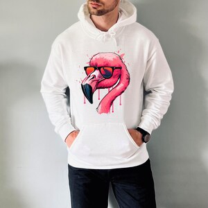 May include: White hoodie featuring a pink flamingo wearing sunglasses. The flamingo design has a black beak and is set against a splatter effect. The hoodie has a front pocket and a drawstring hood.