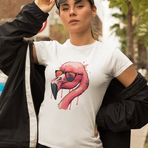 May include: White t-shirt featuring a pink flamingo wearing sunglasses. The flamingo has a pink and red color scheme with dripping accents. The model is wearing black sunglasses and a black jacket.