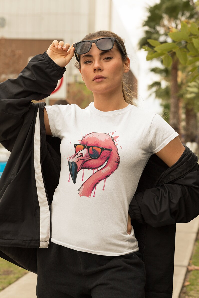 May include: White t-shirt featuring a pink flamingo wearing sunglasses. The flamingo has a pink and red color scheme with dripping accents. The model is wearing black sunglasses and a black jacket.