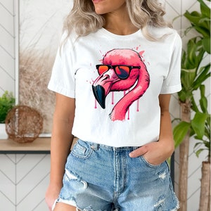 May include: White t-shirt featuring a pink flamingo wearing sunglasses. The flamingo has a black beak and pink dripping accents. The shirt is paired with denim shorts.
