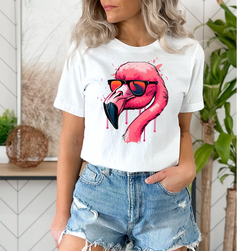 May include: White t-shirt featuring a pink flamingo wearing sunglasses. The flamingo has a black beak and pink dripping accents. The shirt is paired with denim shorts.