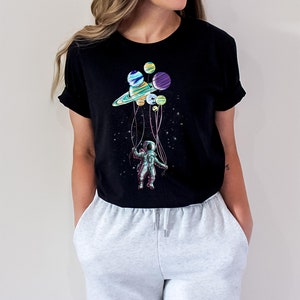 Peut inclure: T-shirt noir avec un graphisme color&eacute; d'un astronaute tenant des ballons en forme de plan&egrave;tes. Le motif comprend une plan&egrave;te semblable &agrave; Saturne et d'autres corps c&eacute;lestes, avec l'astronaute suspendu dans un espace &eacute;toil&eacute;.