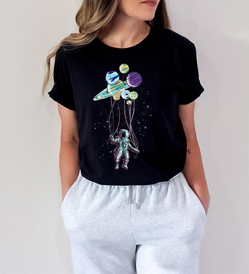 Peut inclure: T-shirt noir avec un graphisme color&eacute; d'un astronaute tenant des ballons en forme de plan&egrave;tes. Le motif comprend une plan&egrave;te semblable &agrave; Saturne et d'autres corps c&eacute;lestes, avec l'astronaute suspendu dans un espace &eacute;toil&eacute;.