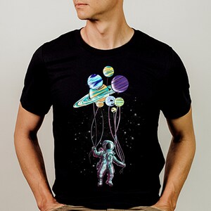 Peut inclure: T-shirt noir avec un motif color&eacute; d'un astronaute tenant des ballons en forme de plan&egrave;tes. Les plan&egrave;tes sont de diff&eacute;rentes couleurs, dont le bleu, le violet et le jaune. L'astronaute porte une combinaison spatiale, sur fond de nuit &eacute;toil&eacute;e.