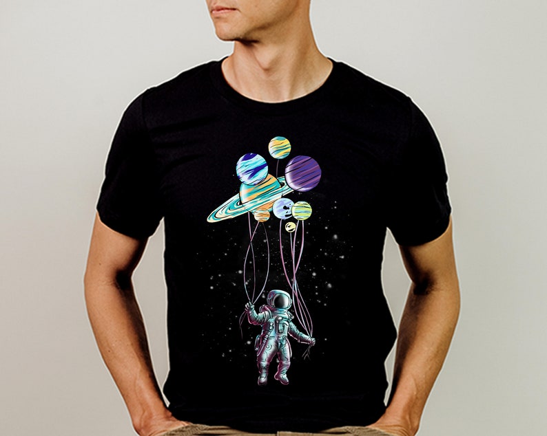 Peut inclure: T-shirt noir avec un motif color&eacute; d'un astronaute tenant des ballons en forme de plan&egrave;tes. Les plan&egrave;tes sont de diff&eacute;rentes couleurs, dont le bleu, le violet et le jaune. L'astronaute porte une combinaison spatiale, sur fond de nuit &eacute;toil&eacute;e.