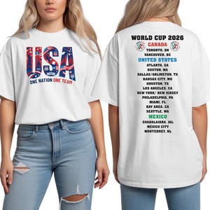 A white t-shirt with text and graphics related to the World Cup, featuring the text "USA" and a list of cities in the United States.