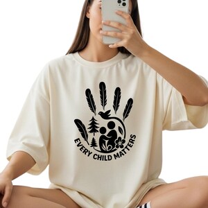 A woman is sitting down and holding a smartphone up to her face, wearing a white t-shirt with a graphic design and text.
