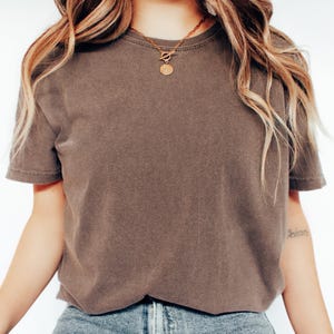 May include: A short-sleeved, brown t-shirt with a crew neck. A gold-colored necklace with a circular pendant is worn. The model is wearing blue jeans. The shirt has a slightly faded appearance.