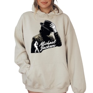 May include: A beige hooded sweatshirt featuring a graphic print of Michael Jackson in a black fedora and sequined jacket. The design includes a silhouette of the artist in a dance pose and the text Michael Jackson in a white script font.