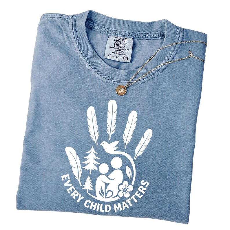 A blue t-shirt with a white graphic design that says "EVERY CHILD MATTERS" and features a handprint, feathers, a bird, a pine tree, and a flower.