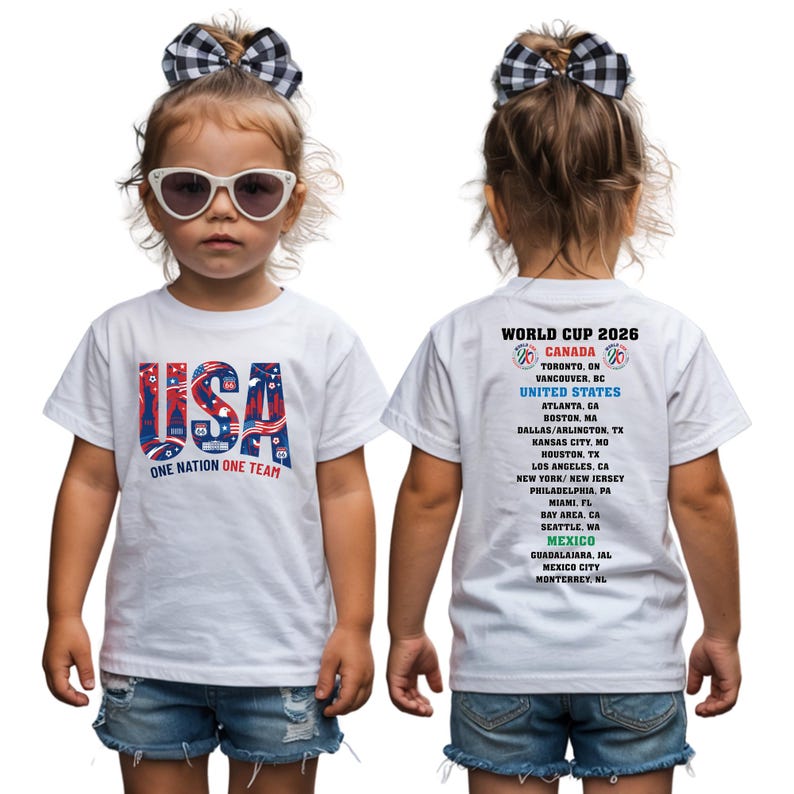 A young girl wearing a white t-shirt with the text "USA ONE NATION ONE TEAM" printed on it, along with a bow in her hair.