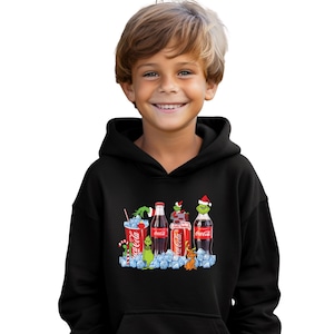 May include: Black hooded sweatshirt featuring a graphic of Coca-Cola bottles and a Grinch-themed design. The design includes a Coca-Cola cup with a candy cane, bottles, and the Grinch character. The sweatshirt is worn by a child.