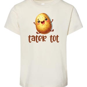 May include: Cream-colored t-shirt featuring a cartoon potato with a smiling face and outstretched arms. The words "tater tot" are printed in brown below the potato illustration. The shirt is made of a soft, comfortable material.