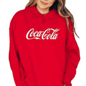 May include: A red hooded sweatshirt with the Coca-Cola logo in white script across the chest. The sweatshirt has a drawstring hood and long sleeves. The classic Coca-Cola logo is a recognizable design.