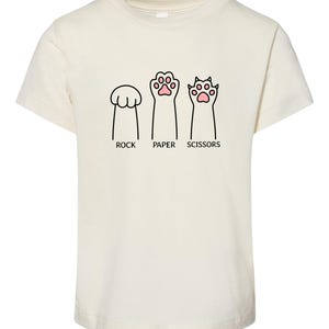 May include: Cream-colored t-shirt featuring a playful design of cat paws representing Rock, Paper, and Scissors. The text "ROCK", "PAPER", and "SCISSORS" is printed below each paw illustration. A fun, casual tee.