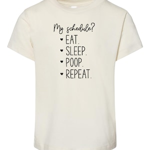 May include: A white t-shirt with black text that reads "My schedule? • EAT. • SLEEP. • POOP. • REPEAT."