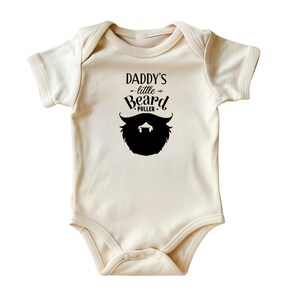 May include: A cream-colored baby onesie with black text that reads "Daddy's little Beard Puller" and a black silhouette of a beard.