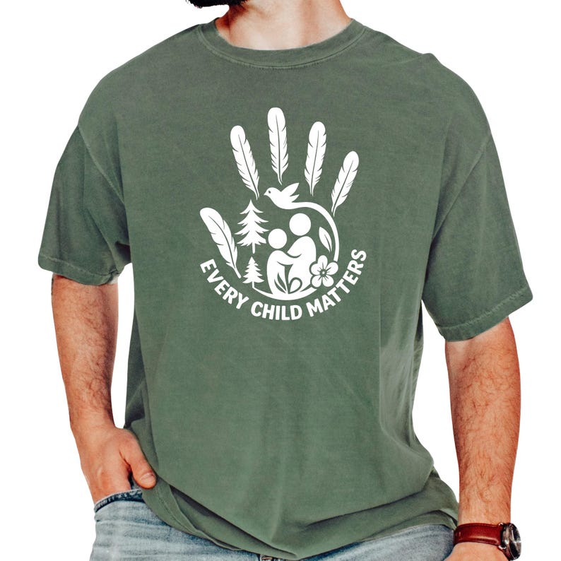 A man wearing a green t-shirt with a white graphic design that says "EVERY CHILD MATTERS" and features a handprint with feathers and a flower.