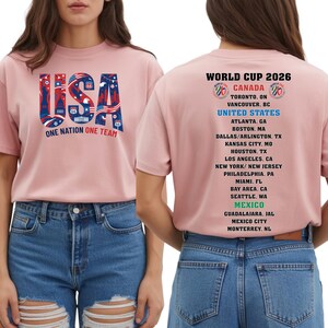 A woman wearing a pink t-shirt with the text "USA" printed on it, along with a list of cities and locations associated with the World Cup.
