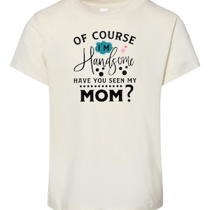 May include: Cream-colored t-shirt with the text "OF COURSE I'M Handsome HAVE YOU SEEN MY MOM?" in black script and block letters. The design includes a blue cloud and pink star graphic.