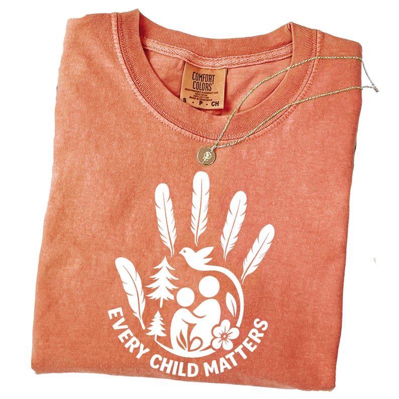 a red t-shirt with a white graphic design on it, which appears to be a handprint with feathers and a flower. The text "EVERY CHILD MATTERS" is printed below the handprint design.