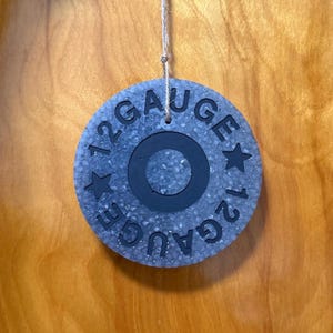 May include: A circular gray textured ornament hanging from a thin string. The surface features a black center ring and the embossed black text 12 GAUGE with two stars. The design mimics the base of a shotgun shell, presented in a flat, round form.