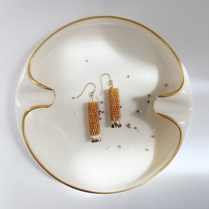 May include: A pair of gold, white, and brown beaded earrings on a white ceramic dish with a gold rim.