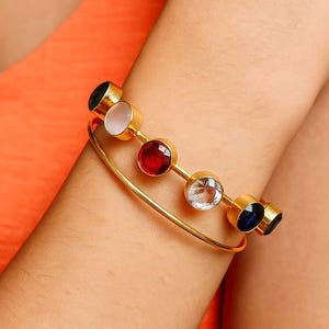 Könnte beinhalten: Goldener Armreif mit fünf farbigen Edelsteinen: Schwarz, Weiß, Rot, Klar und Blau. Die runden Edelsteine sind in einzelnen goldenen Fassungen gefasst, was ein elegantes Accessoire ergibt.