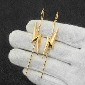 Op de afbeelding: Een paar goudkleurige bliksemschicht oorbellen. Elke oorbel heeft een gestileerde bliksemschicht hanger die is opgehangen aan een dunne, langwerpige haak. De oorbellen worden gepresenteerd tegen een witte achtergrond.
