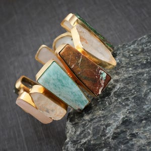 May include: A cuff bracelet featuring multiple raw, irregularly shaped stones in shades of light pink, translucent white, turquoise, and brown, each set in a gold-toned metal frame. The stones are arranged in a row, showcasing varied textures.
