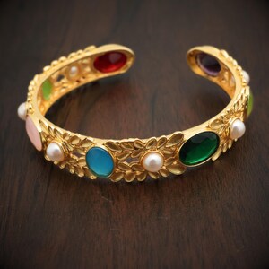 Könnte beinhalten: Goldfarbenes Armreif-Armband mit farbigen Edelsteinen und Kunstperlen. Das offene Armband hat ovale rote, blaue, grüne und violette Edelsteine sowie rosa und grüne Steine und kleine Perlen. Das Armband hat ein florales Design.