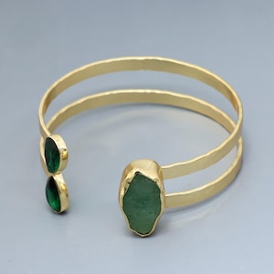 May include: A gold-toned open cuff bracelet featuring a hammered metal finish with two bands. One end is adorned with two small, oval-shaped dark green stones, while the other end features a single, larger, irregular-shaped light green stone.