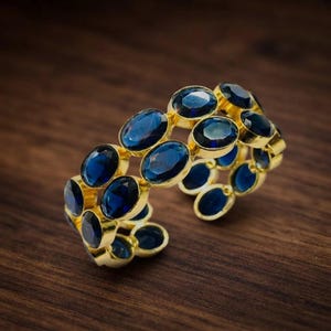 Peut inclure: Un bracelet manchette doré composé de deux rangées parallèles de pierres ovales facettées bleu foncé. Le design ouvert présente une monture métallique jaune qui maintient chaque pierre précieuse dans un sertissage sécurisé et régulier.