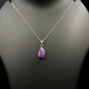 Puede incluir: Un collar de cadena dorada con un colgante de piedra preciosa p&uacute;rpura en bruto de forma irregular. La piedra preciosa tiene una mezcla de morado intenso y tonos lavanda m&aacute;s claros. El collar se exhibe en un maniqu&iacute; negro.