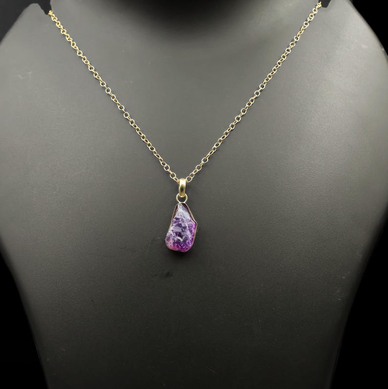 Puede incluir: Un collar de cadena dorada con un colgante de piedra preciosa p&uacute;rpura en bruto de forma irregular. La piedra preciosa tiene una mezcla de morado intenso y tonos lavanda m&aacute;s claros. El collar se exhibe en un maniqu&iacute; negro.
