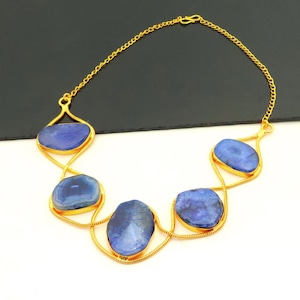 Puede incluir: Un collar dorado con cinco grandes piedras azules irregulares. Las piedras están engarzadas en un delicado marco dorado calado y conectadas por una fina cadena. El collar se muestra sobre un fondo blanco y negro.