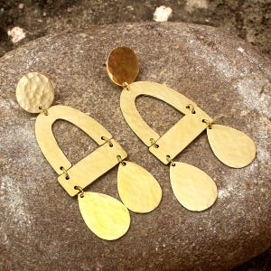 May include: A pair of gold-colored earrings with a hammered texture. Each earring features a circular stud, an arched shape, a rectangular bar, and two teardrop-shaped dangles. The earrings are displayed on a stone surface.