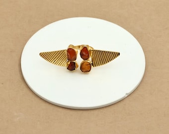 May include: A gold-colored ring with four orange-brown gemstones. The ring has a unique design with two wing-like structures on either side, each with a textured, ridged pattern. The ring is displayed on a white, circular surface.