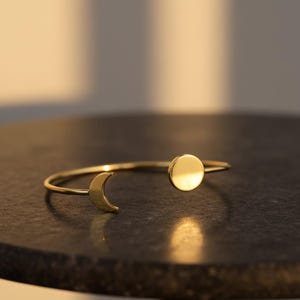 May include: Gold-colored open cuff bracelet featuring a crescent moon and a circular disc design. The bracelet is made of a thin, curved band that wraps around the wrist. The moon and disc are positioned on opposite ends of the bracelet.