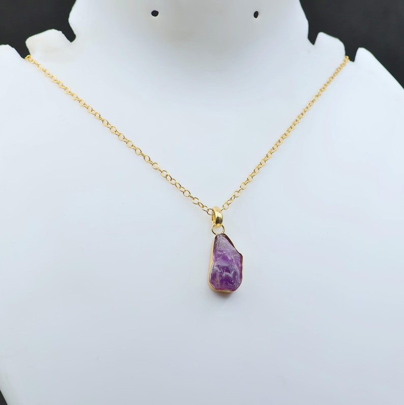 Puede incluir: Un collar de cadena dorada con un colgante de amatista p&uacute;rpura en bruto. La amatista est&aacute; engastada en un bisel dorado. El collar se exhibe en un maniqu&iacute; blanco.