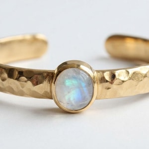 May include: A gold-toned open ring featuring a hammered texture along the band and a single, round, bezel-set moonstone cabochon at the center. The stone displays a translucent white appearance with subtle iridescent blue flashes under the light.