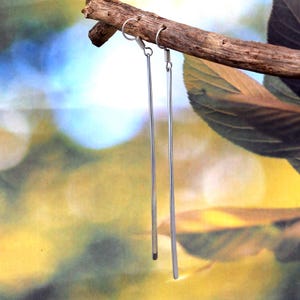 May include: A pair of long, silver-coloured linear earrings suspended from a small wooden branch. Each earring features a simple hook and a straight, rectangular bar. The background is a soft blur of green, yellow, and blue, creating a natural aesthetic.