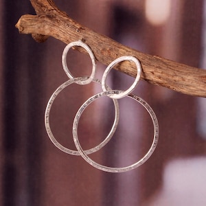 Puede incluir: Pendientes de aro plateados con textura martillada, colgando de una rama de madera. Los pendientes presentan un diseño de círculo abierto grande, con un círculo más pequeño que se conecta al gancho. Los pendientes son de color plateado brillante.