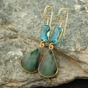 May include: A pair of gold-coloured earrings featuring blue rectangular and green teardrop-shaped gemstones. The earrings are presented on a grey, textured surface. The gemstones have a polished finish and are set in gold-coloured metal.