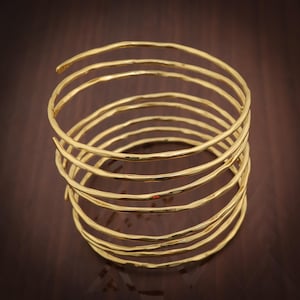 May include: A gold arm cuff with a spiral design. The cuff is made of multiple, textured bands that wrap around the arm. The jewellery piece is displayed on a dark brown surface.