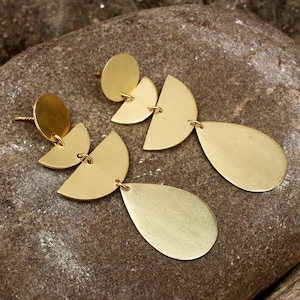 Puede incluir: Un par de pendientes dorados. Cada pendiente presenta un pequeño perno redondo, dos formas de semicírculo y un colgante en forma de lágrima. Los pendientes se exhiben sobre una superficie marrón texturizada.