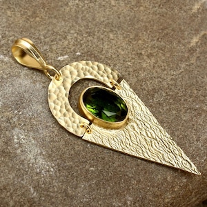 Könnte beinhalten: Goldfarbener Anhänger mit einem grünen, ovalen Edelstein. Der Anhänger hat eine strukturierte, gehämmert Oberfläche auf den kreisförmigen und dreieckigen Elementen. Der Edelstein ist in einer goldenen Fassung gefasst. Für eine Halskette.